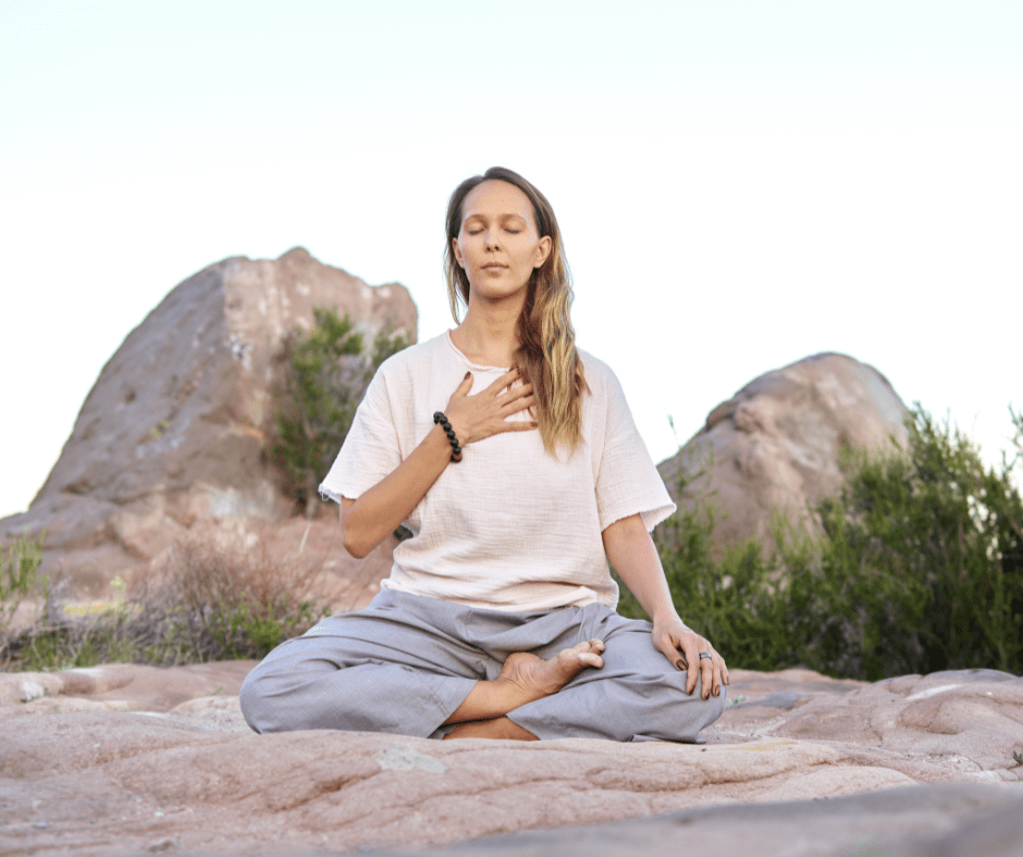 Woman sitting outside near some boulders who is meditating with her hand on her chest and eyes closed. 